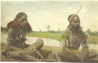229030 Man, vrouw en baby van de Marind-anim zitten aan de oever van een rivier